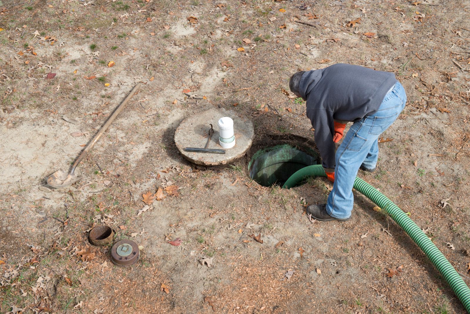 Worker leaning over an open manhole with a green hose on bare ground