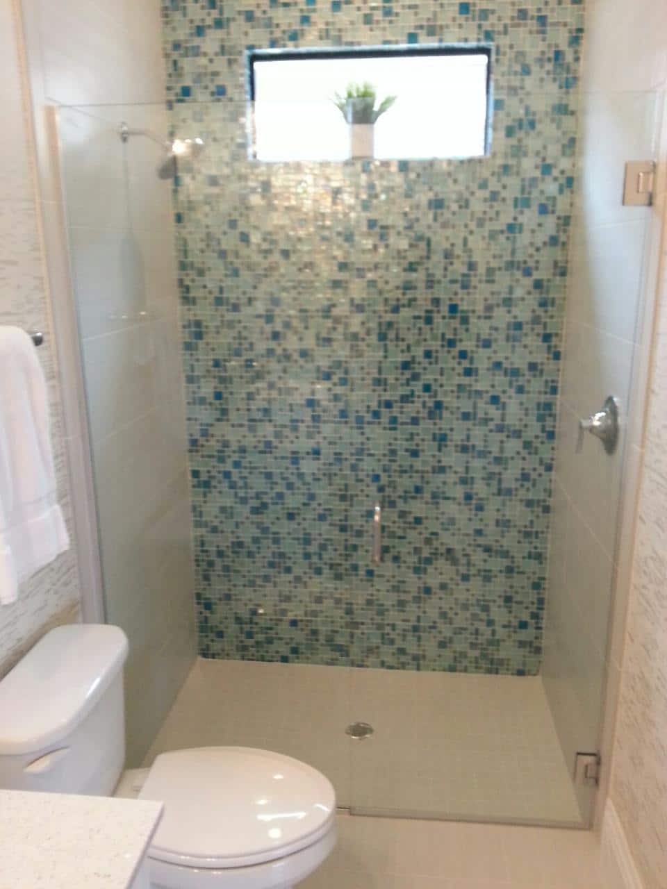 Small modern bathroom with teal tile shower, glass door, toilet, and window with plant.