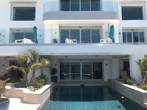 White building with pool, glass balcony, turquoise umbrellas, and palm trees.