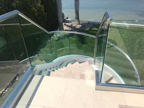Staircase with glass railing, outdoor setting, descending curved stairs, greenery, and water view.