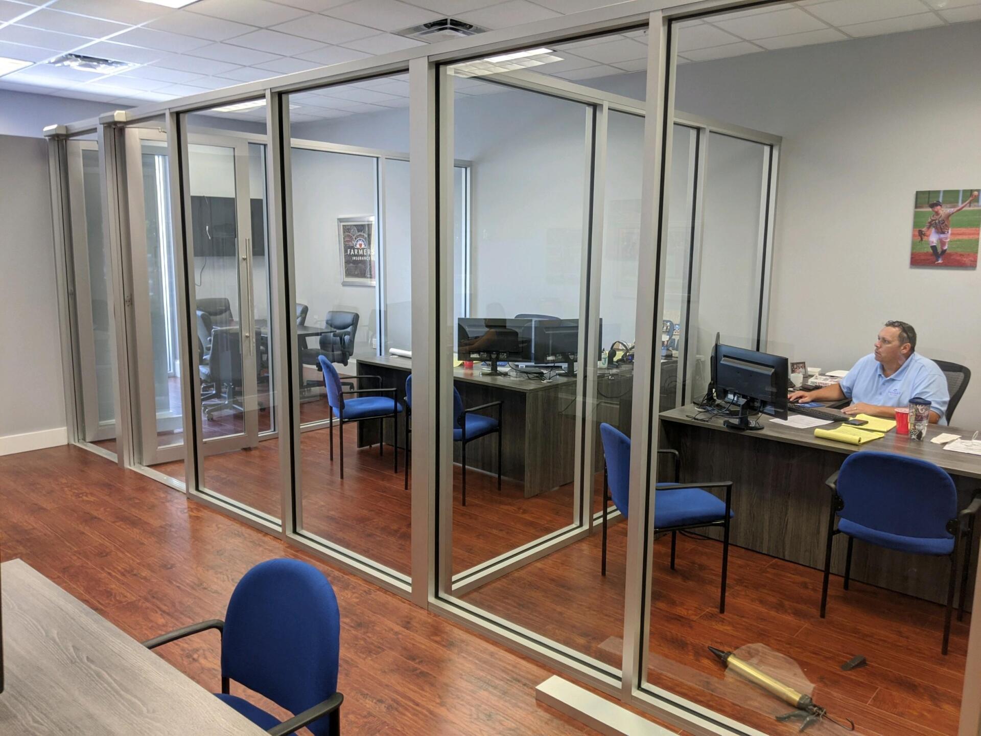Office space with glass walls, wooden floors, desks, computers, and a man working at a desk.