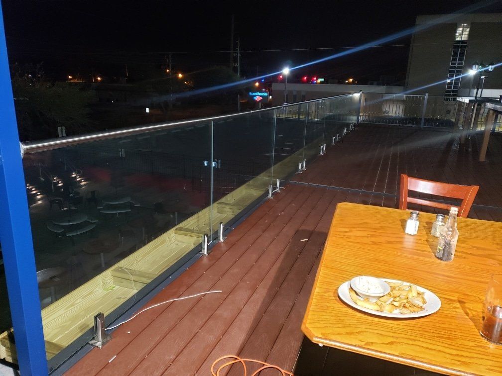 Outdoor restaurant patio at night with glass railing and wooden deck. A table is set with food.