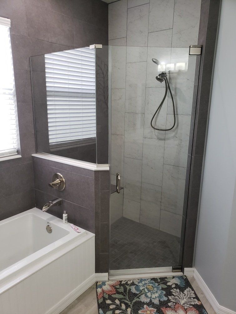 Bathroom with a white tub and a glass-enclosed shower, gray tiles, and a colorful rug.