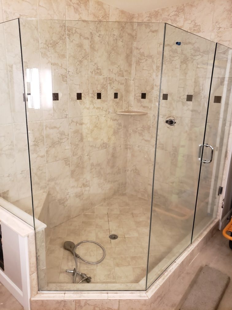 A glass-enclosed corner shower with beige tile walls and floor, a handheld showerhead, and a white cabinet.