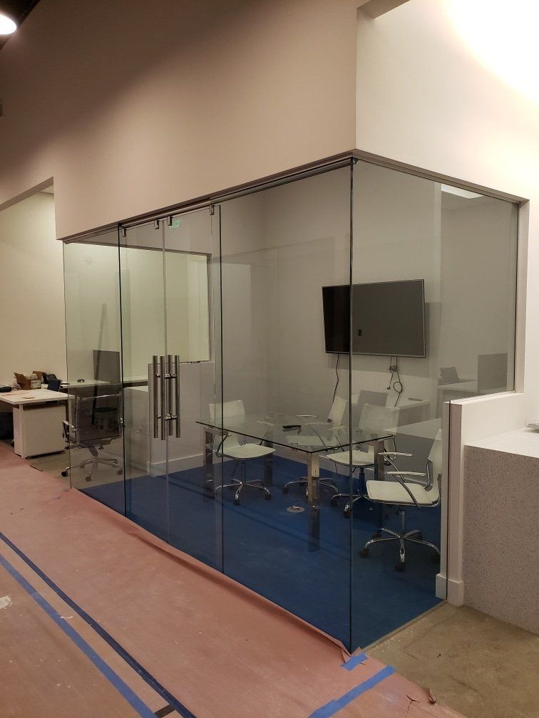 Glass-walled office space with a meeting table and chairs, blue carpet, and a mounted TV.