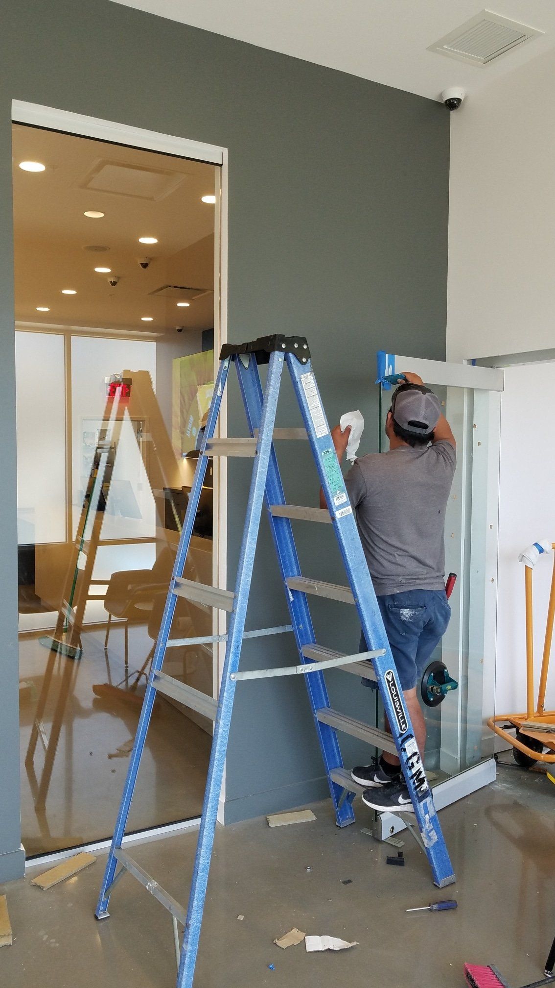 A person on a blue ladder installs something on a gray wall near a glass door.