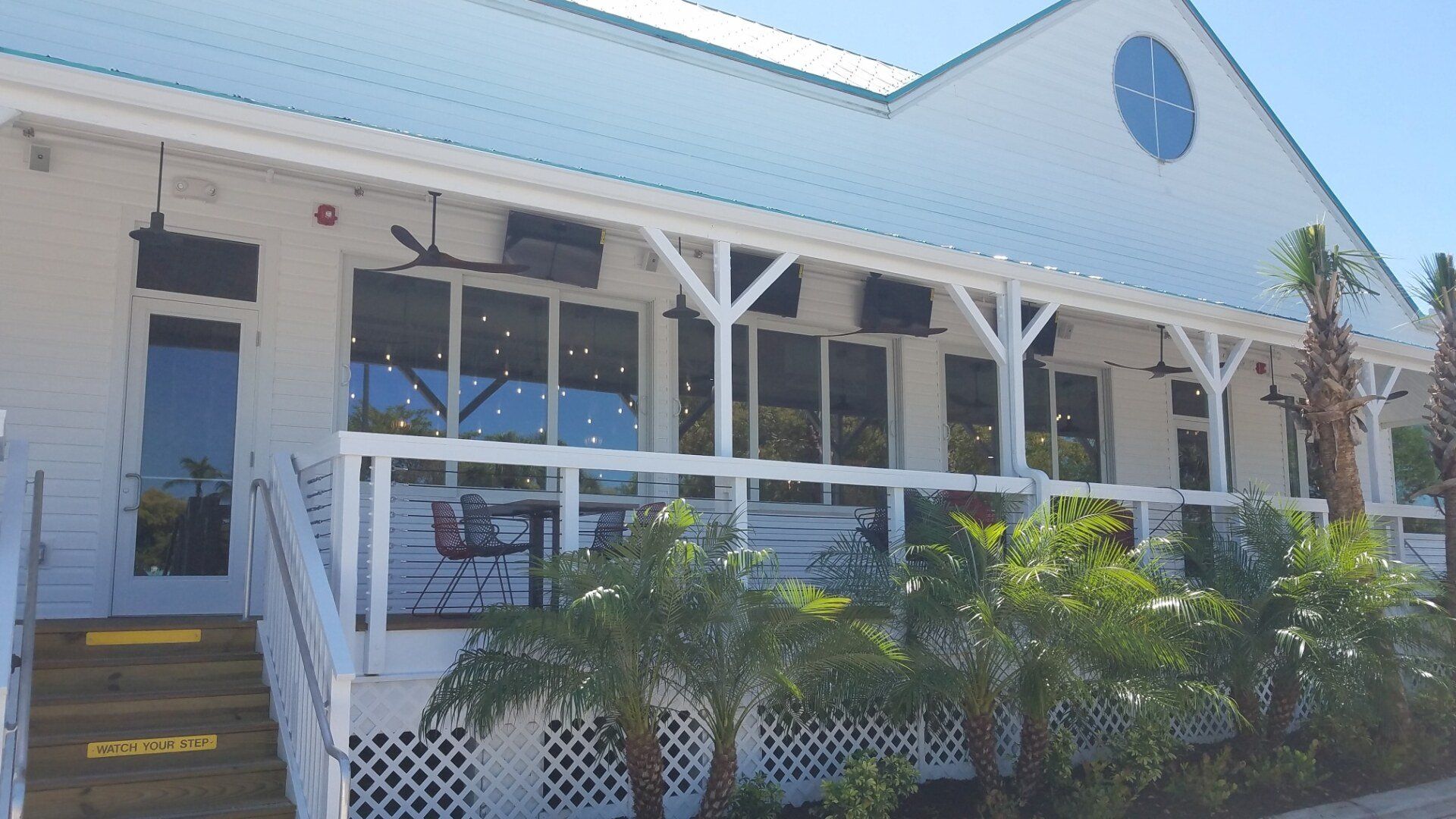 White building with porch and windows, palm trees. Sign says 