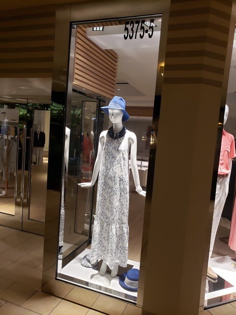 Mannequin in a white dress with blue hat, inside a store window.