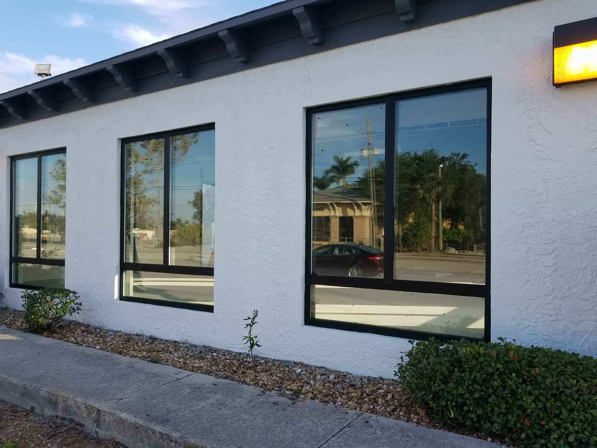 Exterior of a white building with three windows reflecting the street; black frames, gravel, bushes, and a blue sky.