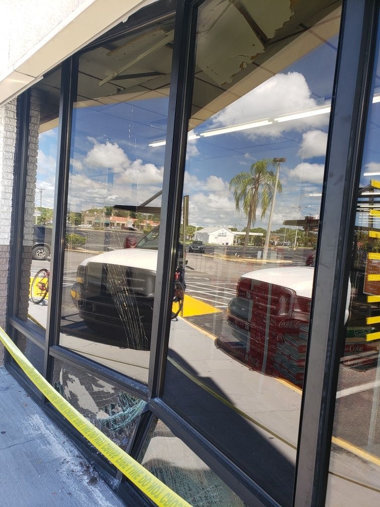 Broken storefront window reflecting a blue sky, clouds, and police car. Yellow caution tape on the ground.