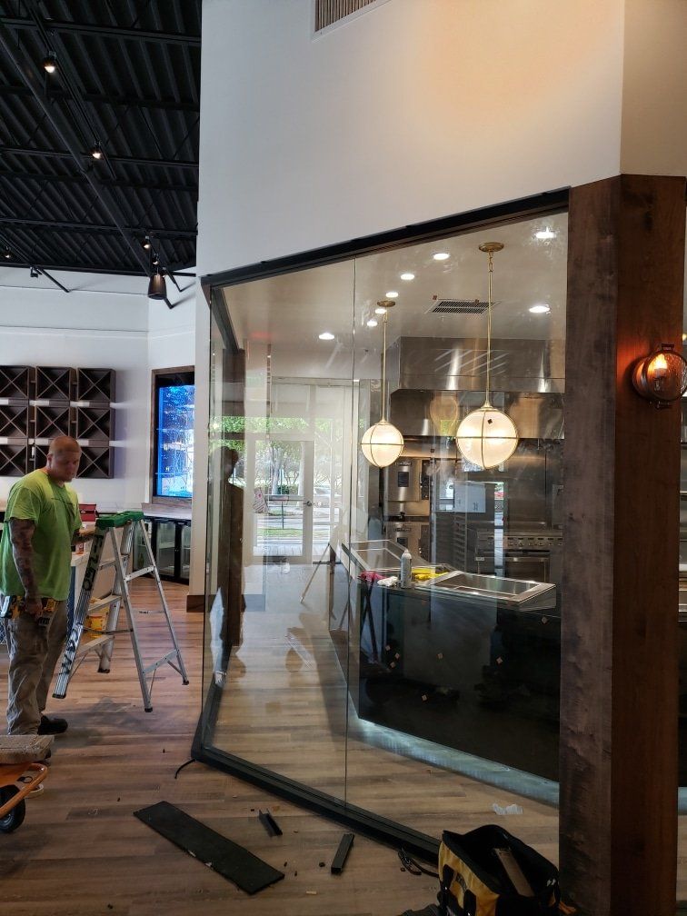 Construction of a restaurant interior with clear glass walls, worker, lights, wood accents, and open kitchen.