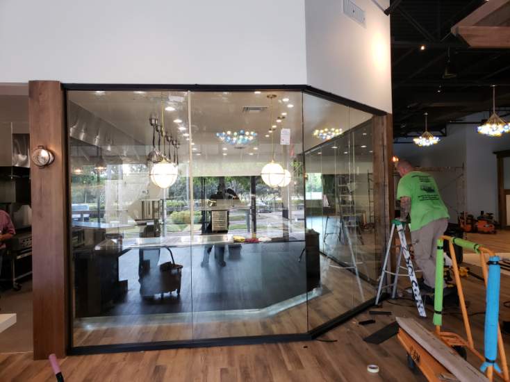 A man on a ladder installs a black-framed glass enclosure inside a commercial space.