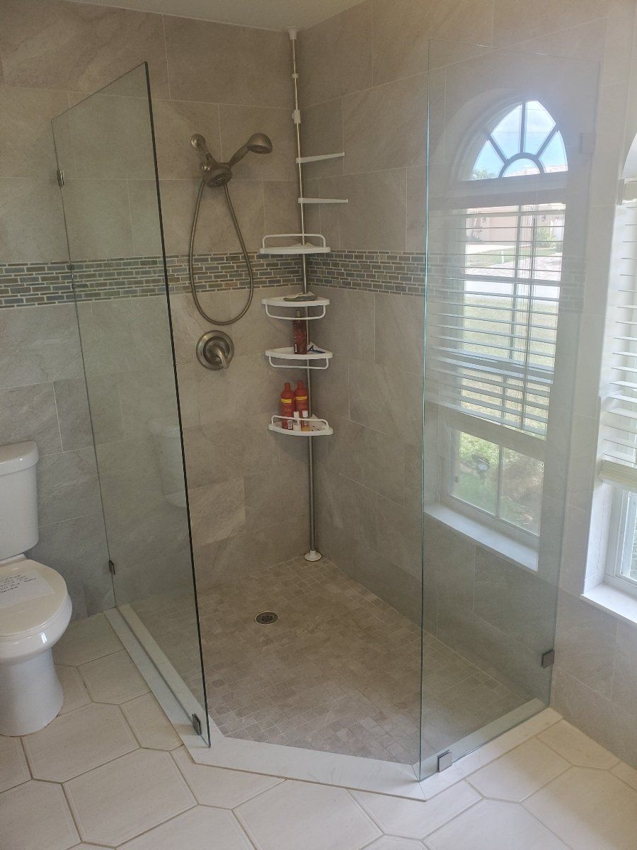 A glass-enclosed shower with a corner window in a bathroom. White and gray tiles. Silver fixtures and shelving.