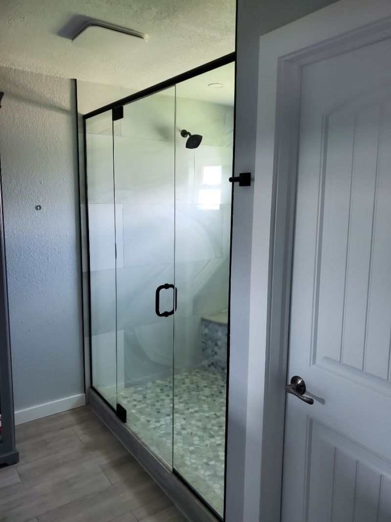Glass-enclosed shower with black framing and door handle, next to a white door. The shower has a stone floor.