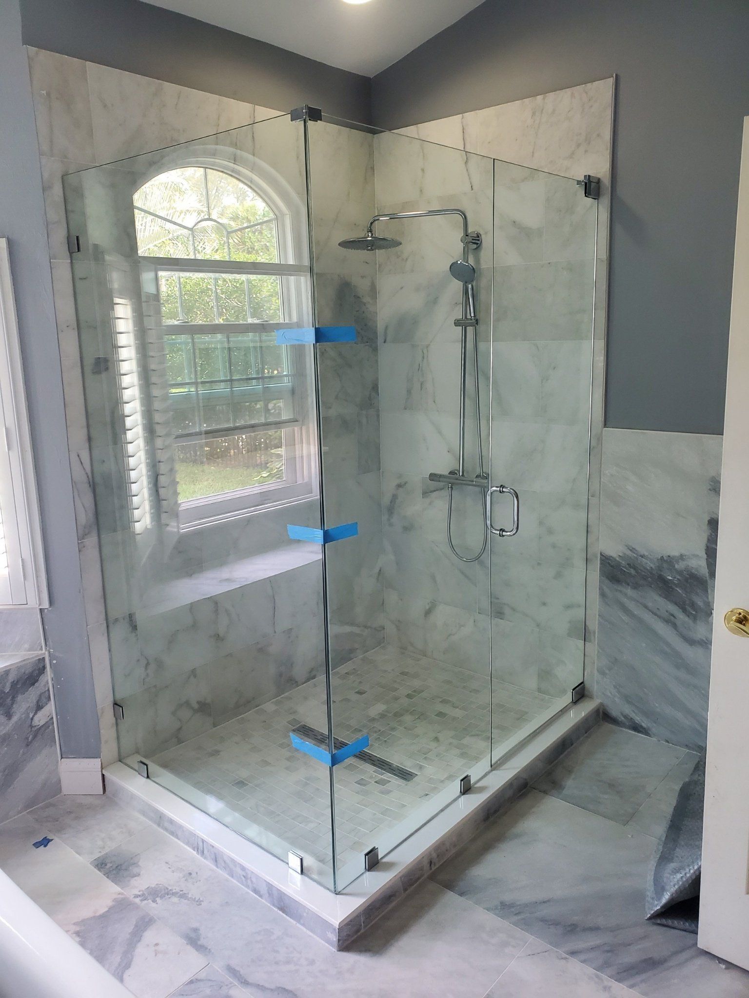 Corner glass shower with marble walls, window, and shower head.