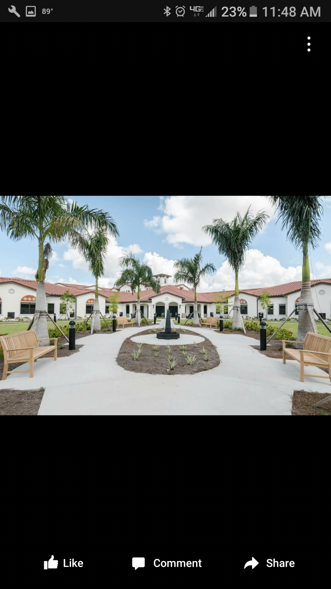 Lush courtyard of a facility, palm trees, fountain, benches, beige building, blue sky.
