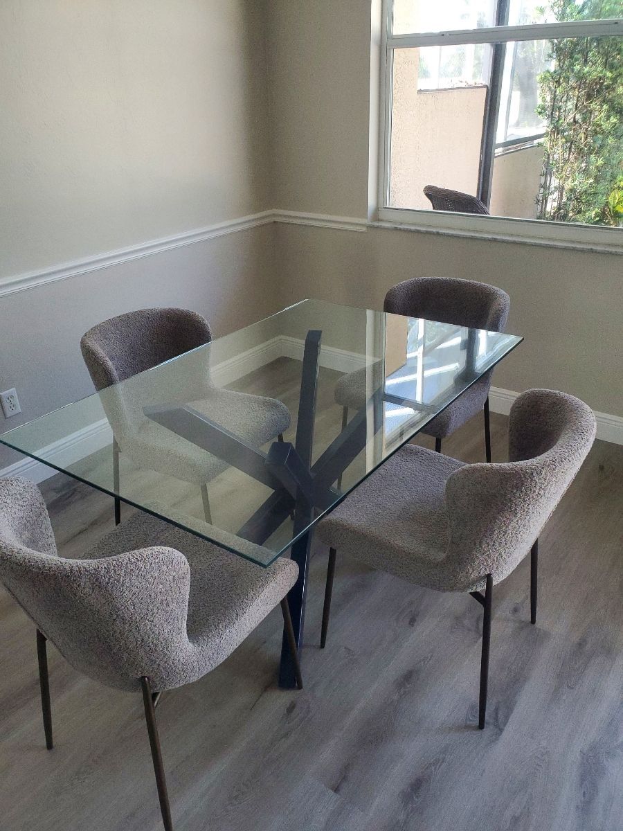 A glass dining table with four gray upholstered chairs. Set against a window and light gray walls.