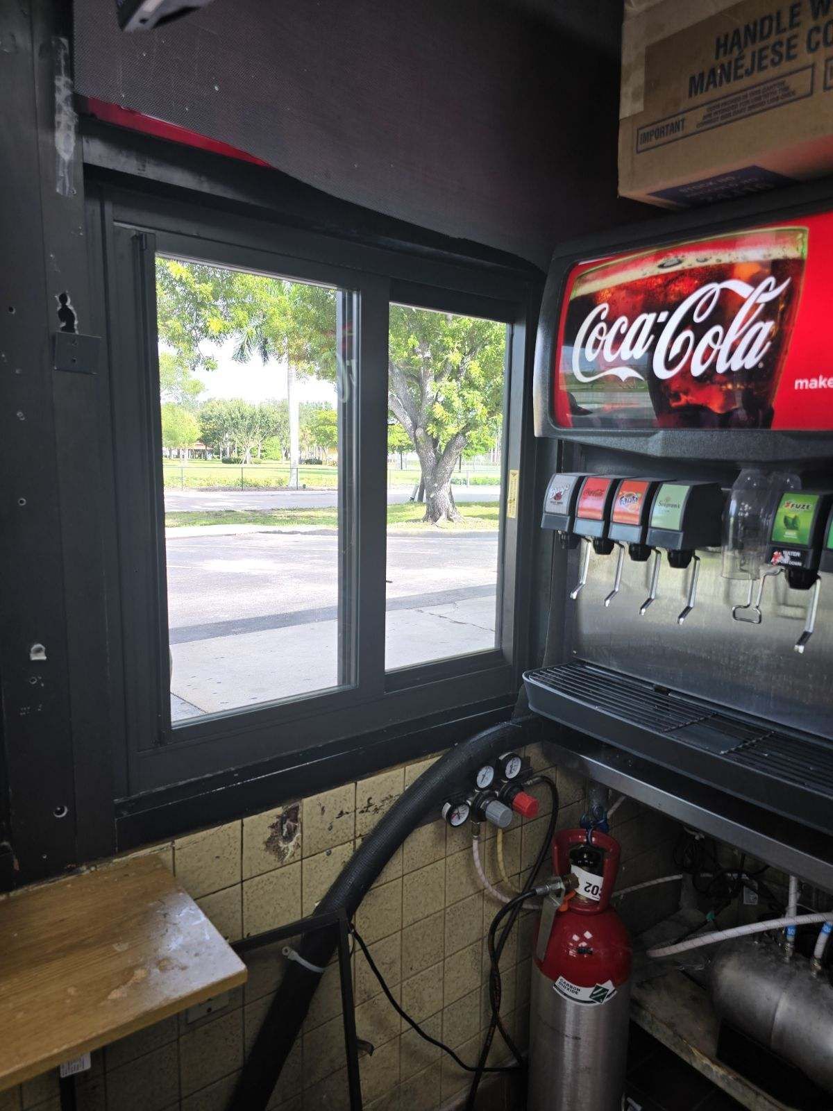 Interior of a restaurant with a Coca-Cola machine, window overlooking a road with trees, and equipment.