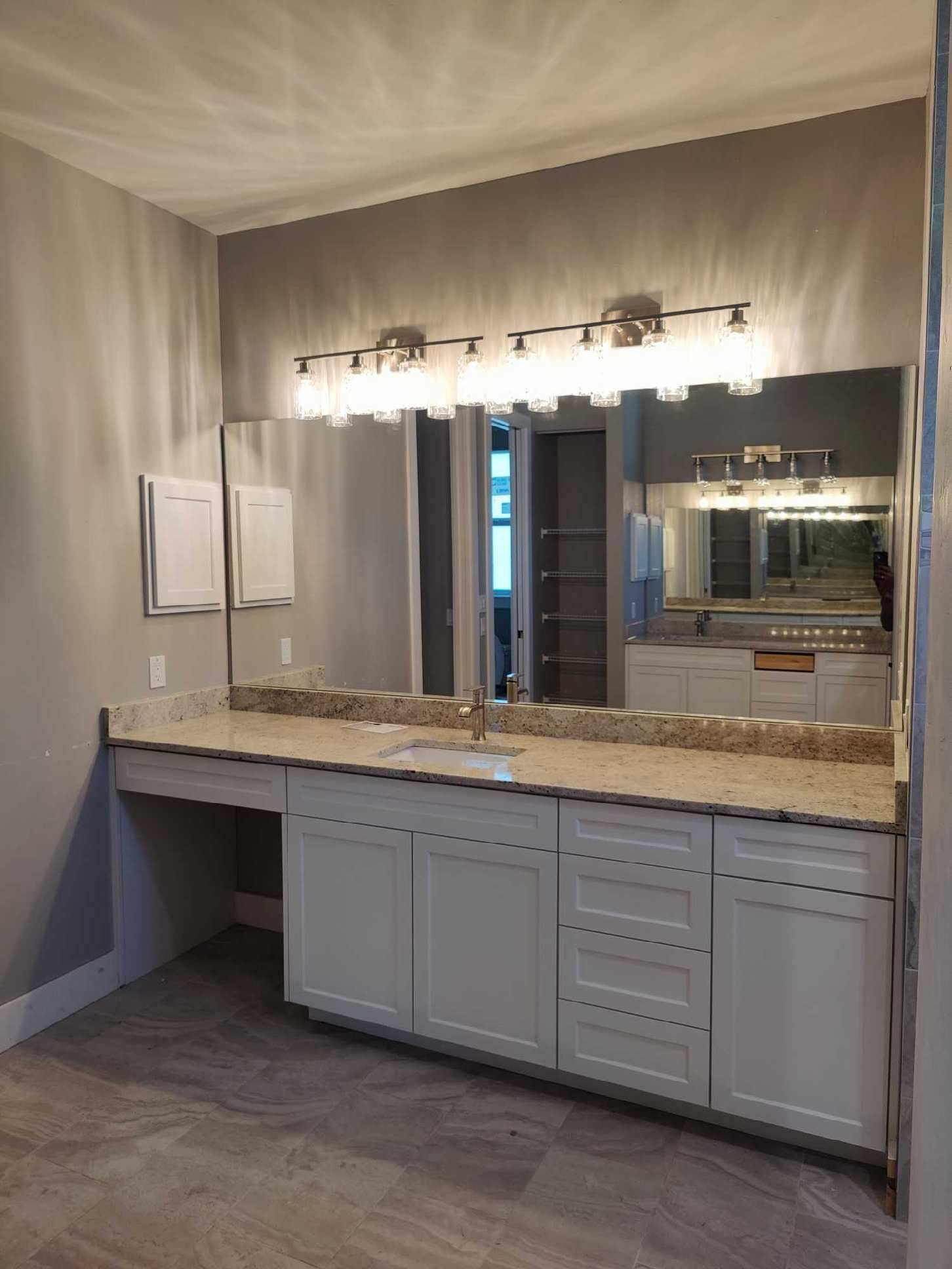 Bathroom with a long countertop, large mirror, white cabinets, and a light fixture. Gray walls and flooring.