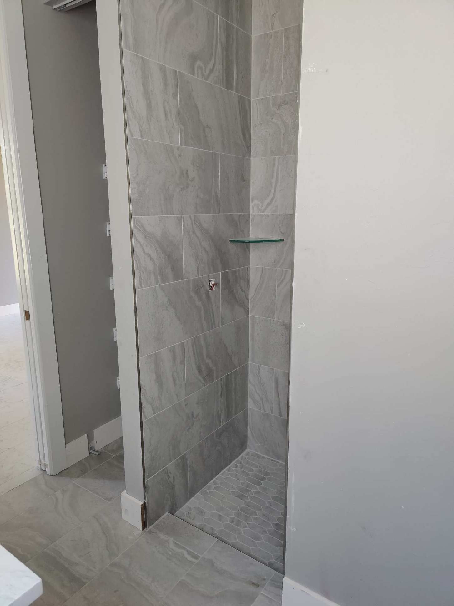 Walk-in shower with gray tiled walls and floor; glass shelf; gray and white walls.