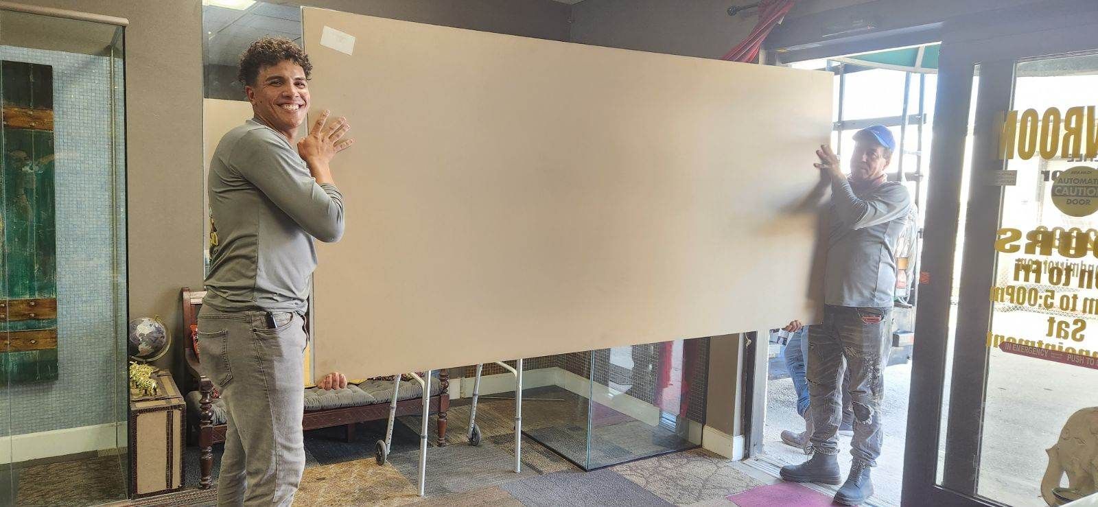 Two people hold a large cardboard sheet in a store doorway. One smiles, clapping.