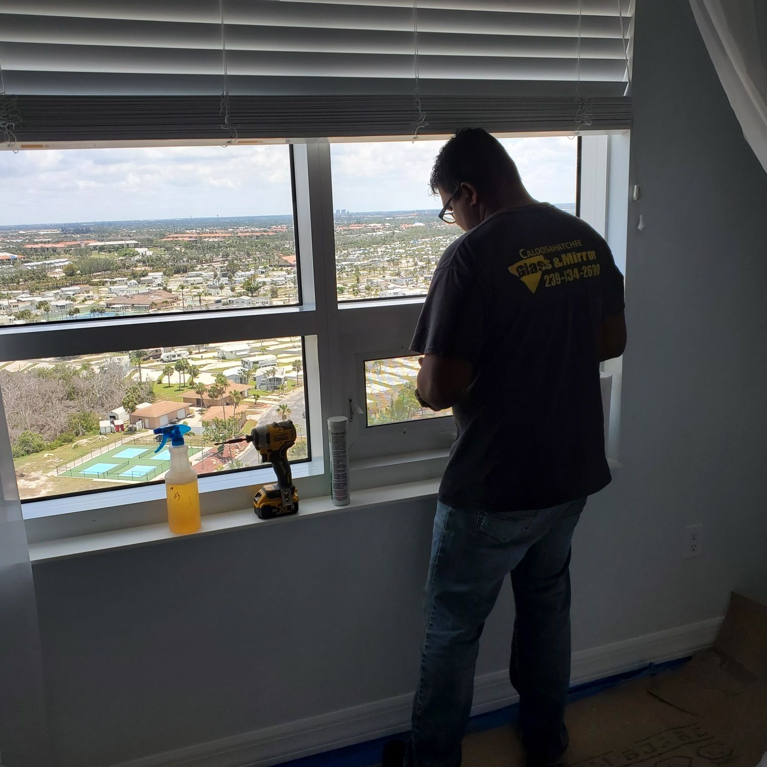A man installs blinds in a bright room with a city view, drill and spray bottle on the window sill.