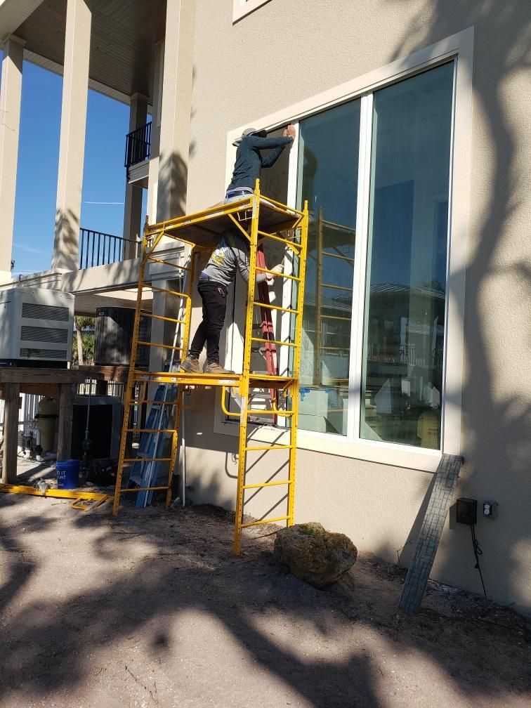 Two people on a yellow scaffold, working on the exterior of a house with large windows.