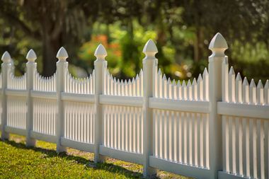 White picket fence with pointed posts along a sunlit lawn, with trees blurred in the background White picket fence with pointed posts along a sunlit lawn, with trees blurred in the background