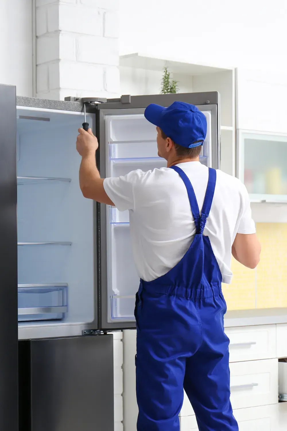 A repairman in blue overalls and cap fixes a refrigerator with a screwdriver in a kitchen.