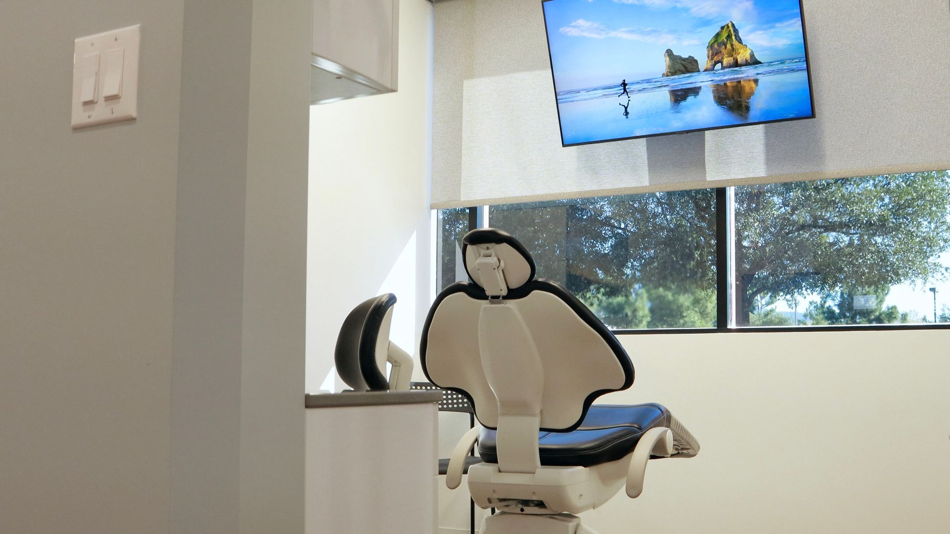 Dental chair in an exam room, with a monitor and window, a light switch on the wall.