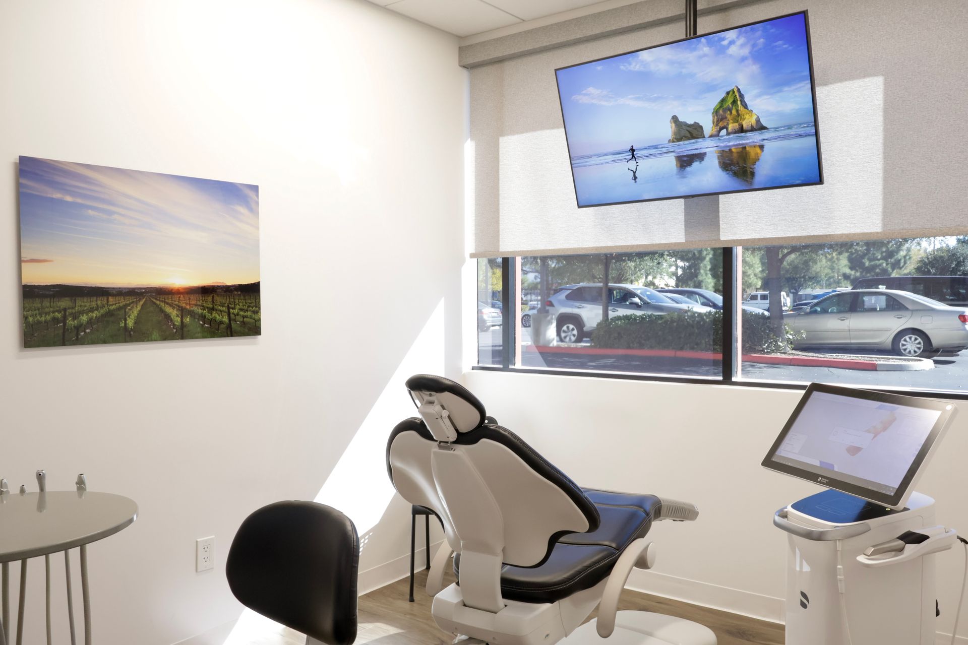 Dental exam room with chair, monitor, window view of cars, and artwork.