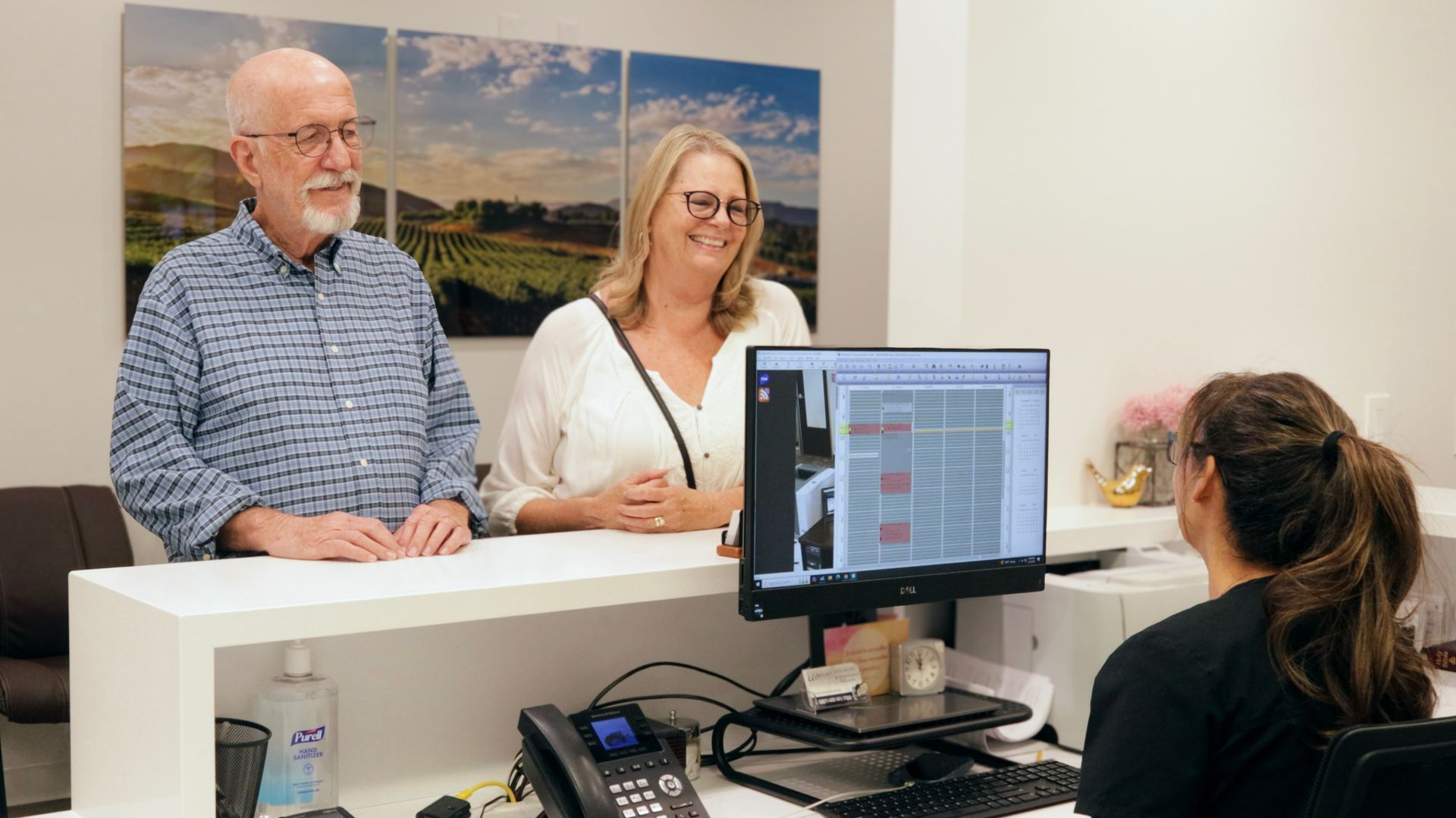 Couple at a reception desk smiles at a staff member, computer screen visible, bright office setting.