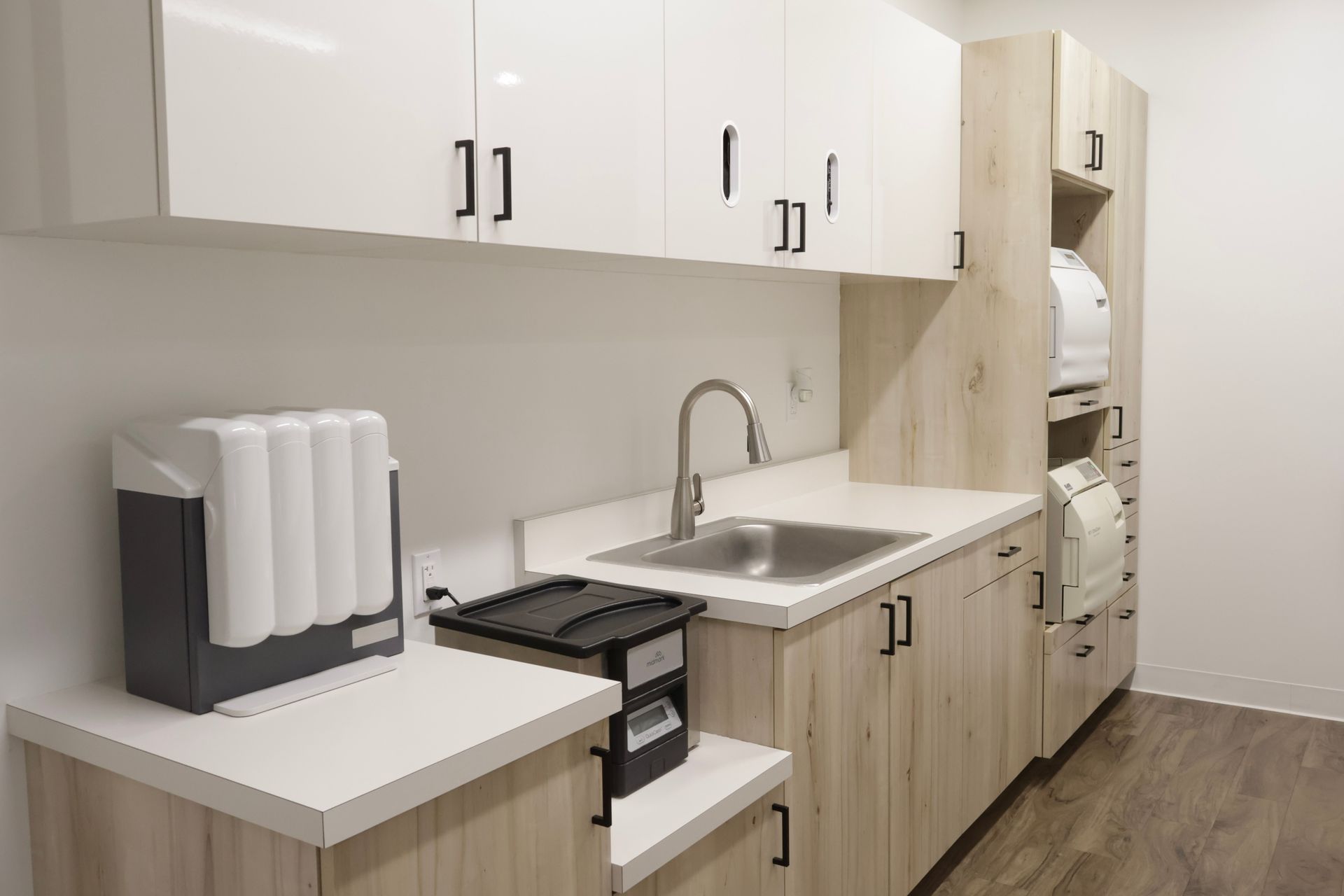 Modern dental office utility room with cabinetry, sink, and equipment. Light wood and white cabinets.