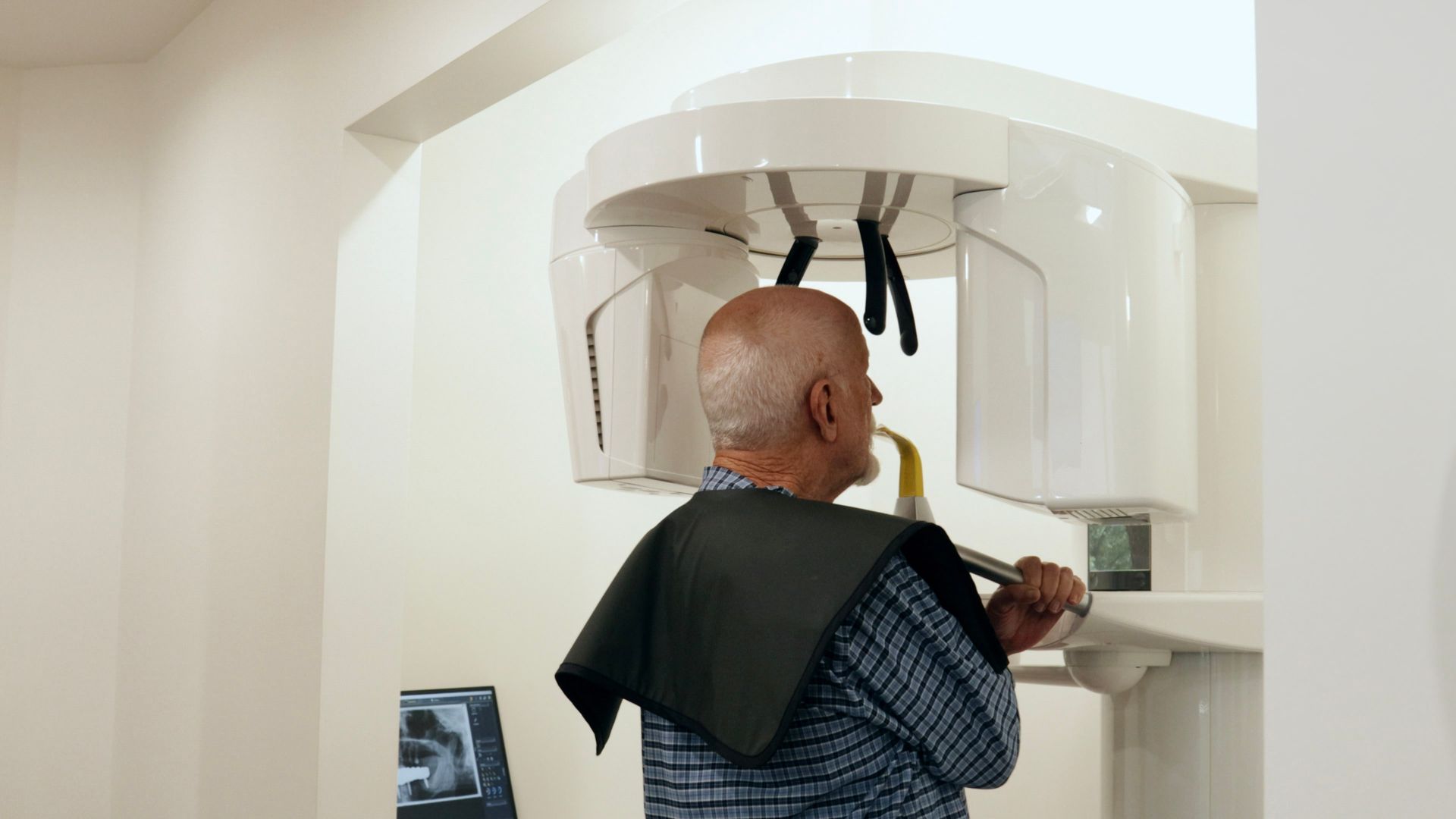 Man standing in dental X-ray machine, wearing protective apron. White dental office.