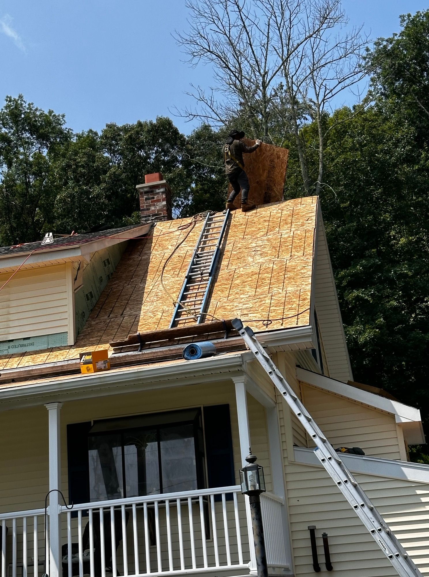 Roofers on a steep roof replacing shingles; house exterior, ladder, chimney, sunny day.
