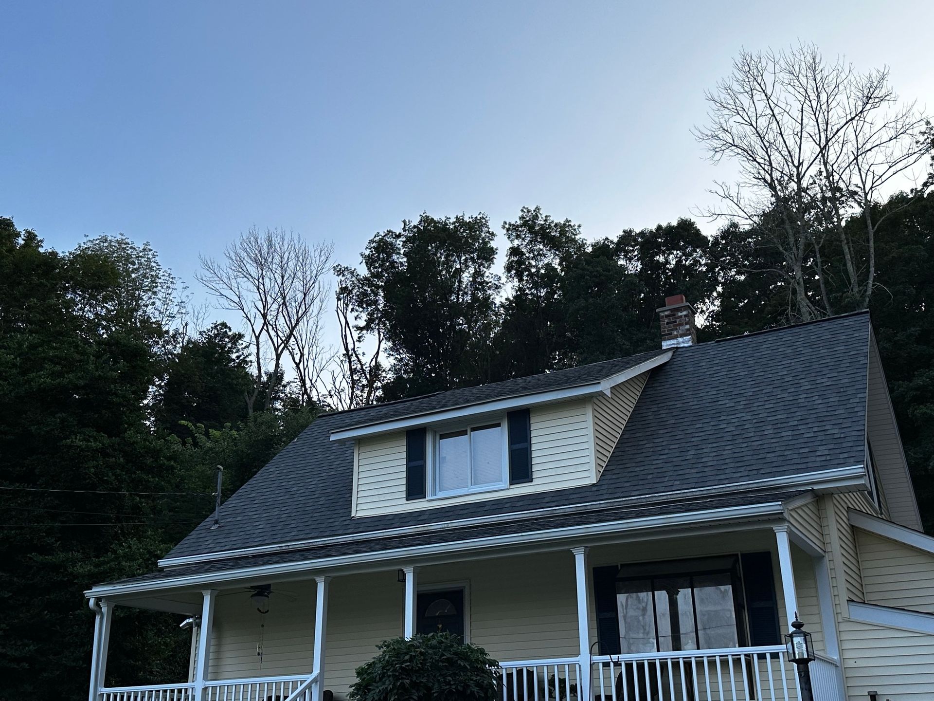 A white house with a black roof and a porch surrounded by trees.
