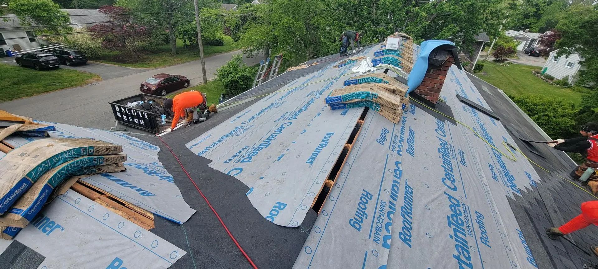 Roofing crew installing gray shingles on a house roof; one worker visible near the ridge, with tools and materials on top.