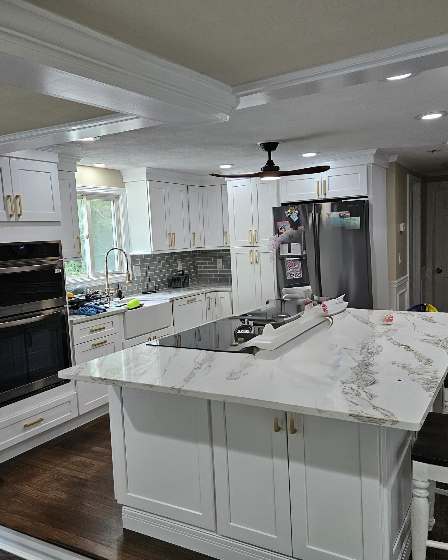 A kitchen with white cabinets , a large island , a stove , a refrigerator and a ceiling fan.