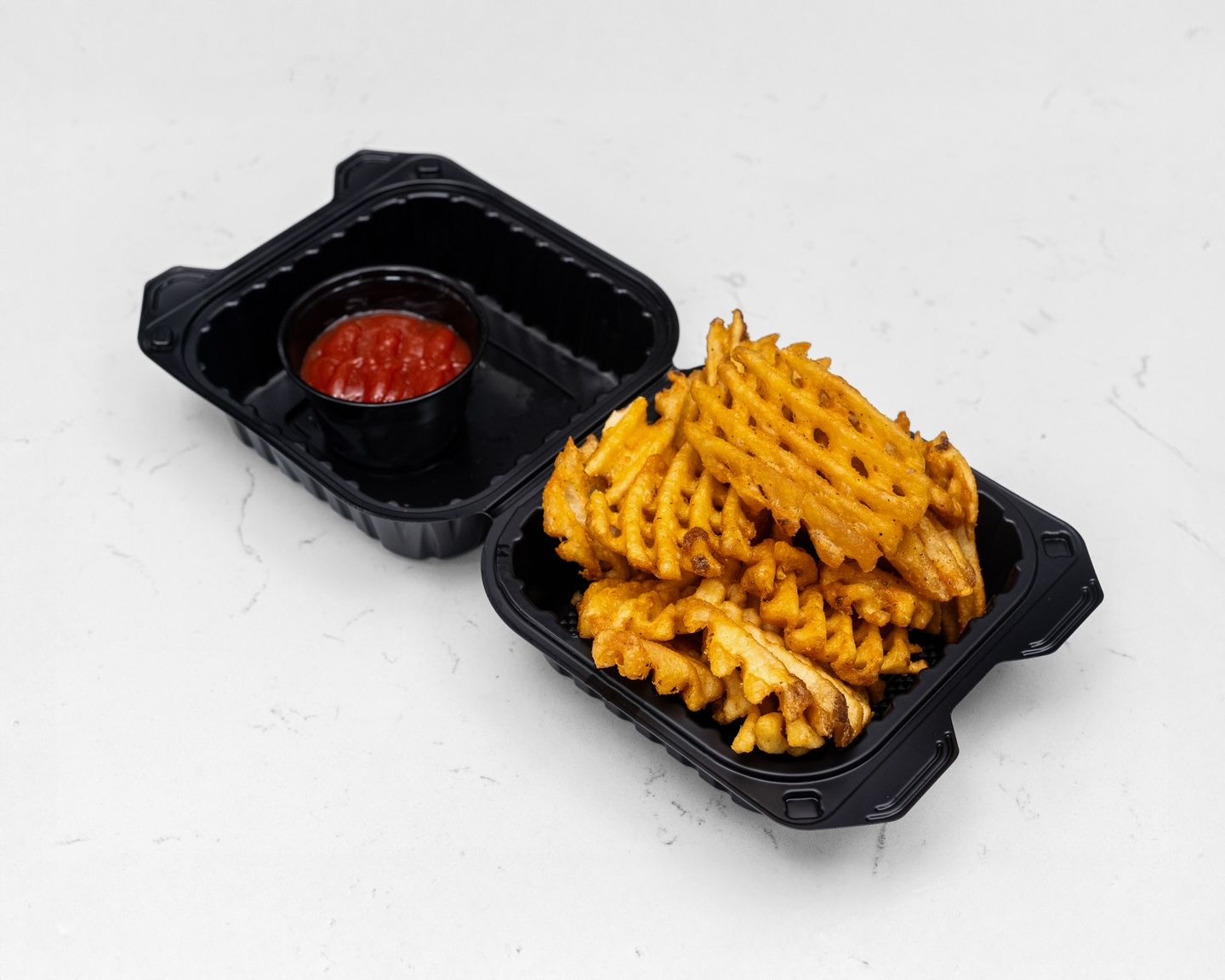 Waffle fries in a black takeout container, ketchup in a separate compartment, on a white surface.
