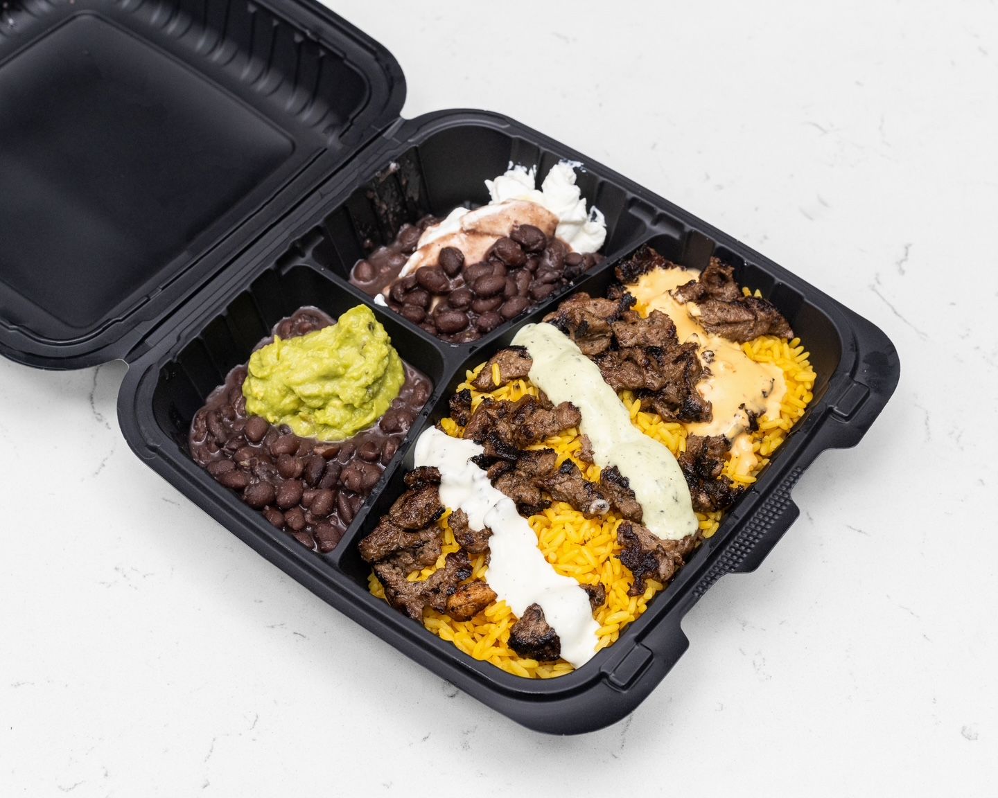 Black takeaway container with rice, meat, beans, guacamole, and sauces.