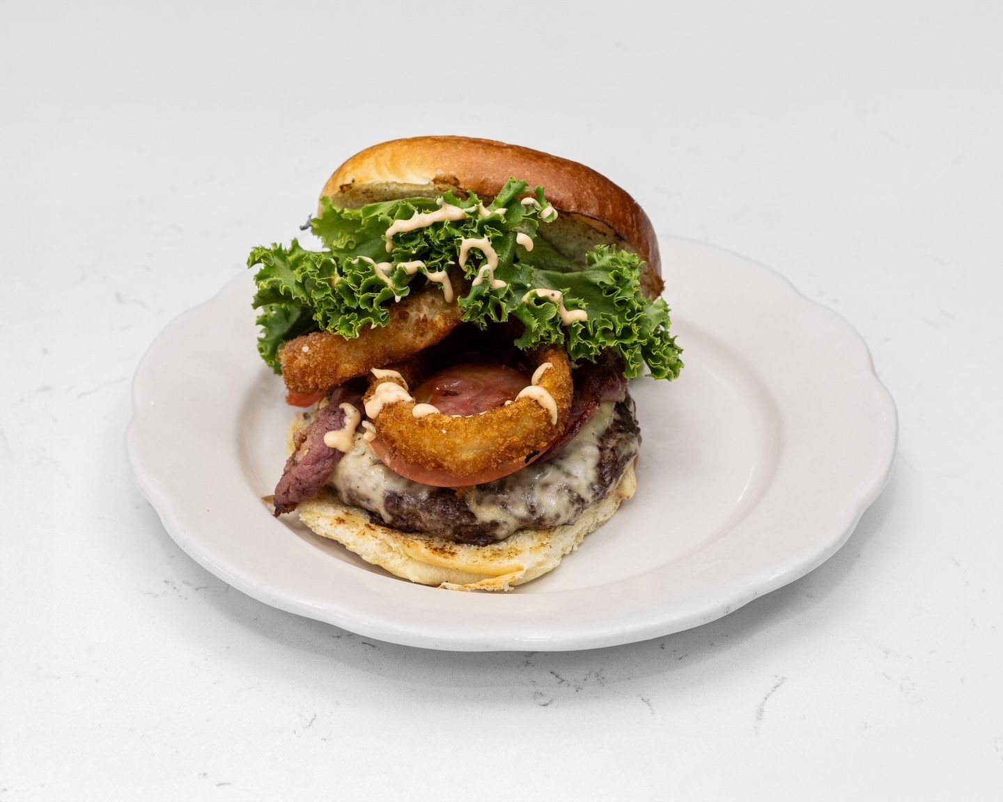 Burger on a plate with onion rings, lettuce, and sauce, against a white background.