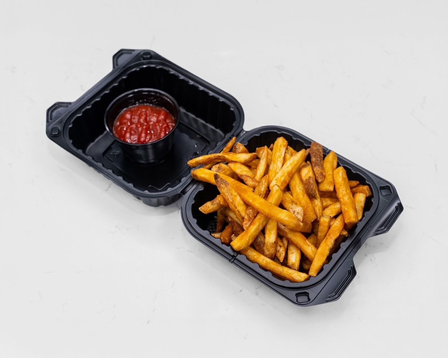 French fries in a black container with a ketchup cup, set on a white surface.