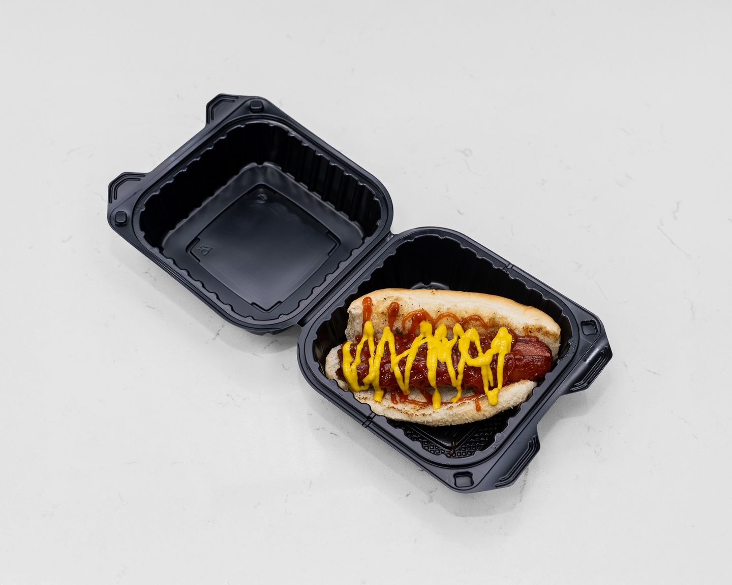 Hot dog in a black plastic container with mustard and sauce on a white surface.