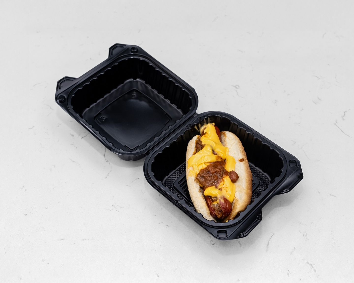 Chili cheese dog in a black takeout container, open on a white surface.