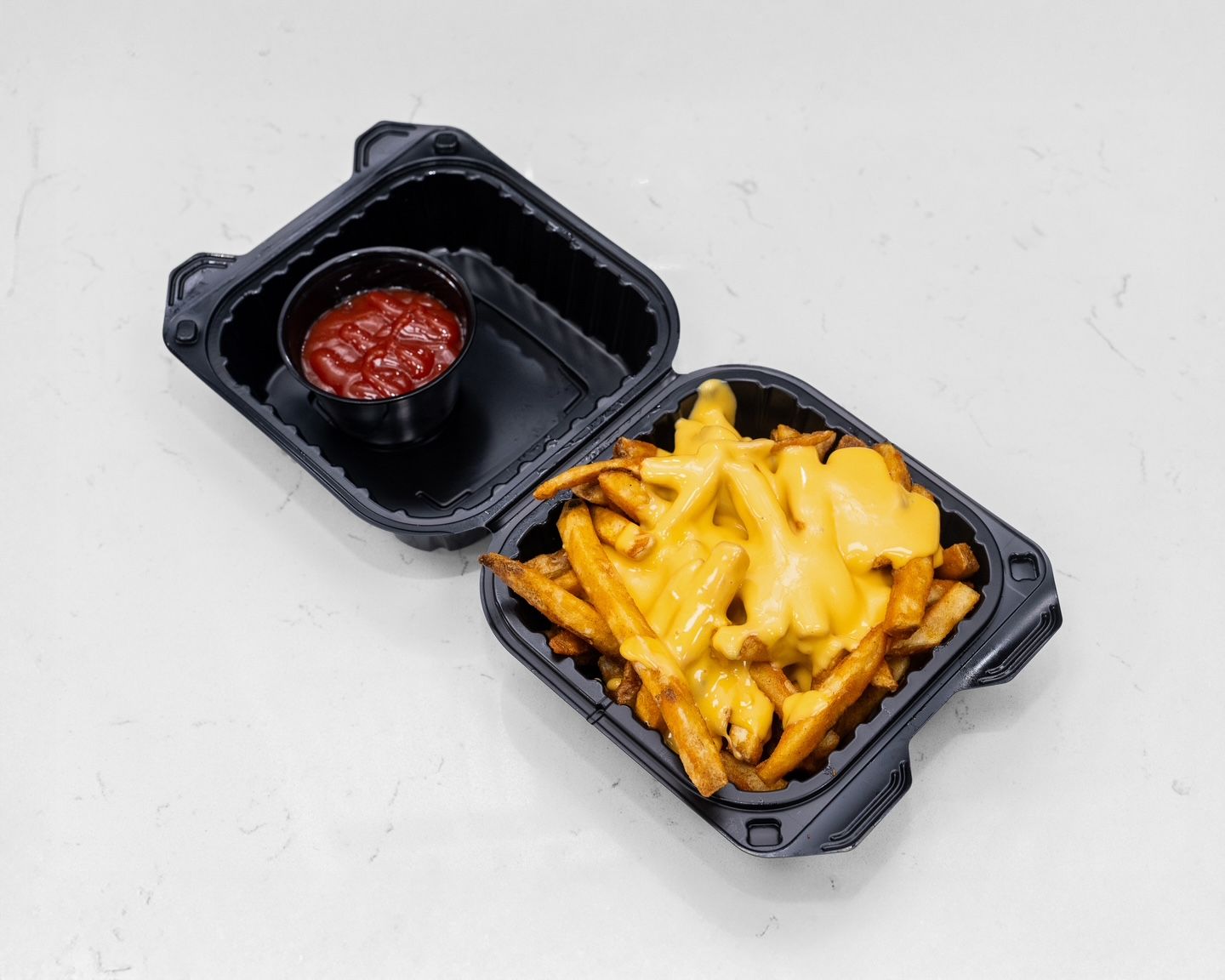 Fries with melted cheese in a black container with ketchup, on a white surface.