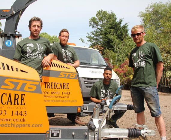 Four tree surgeons from Treecare stand by wood chippers and a van on a sunny day.