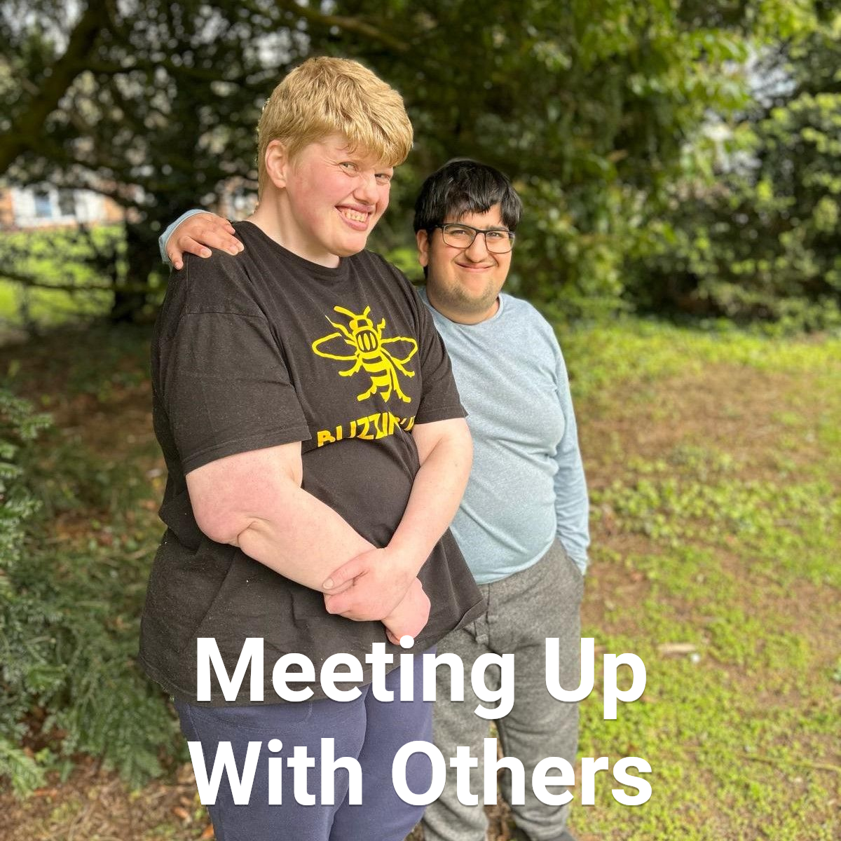 Two smiling people embrace outside. One with blonde hair wearing a black shirt, the other wearing glasses and a blue shirt.