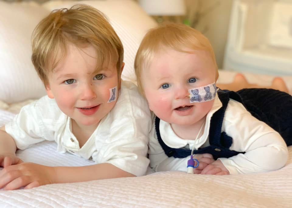 A child and a baby with prader-willi syndrome lying on their fronts smiling.  The baby has a naso-gastric tube covered by a teddy bear plaster.