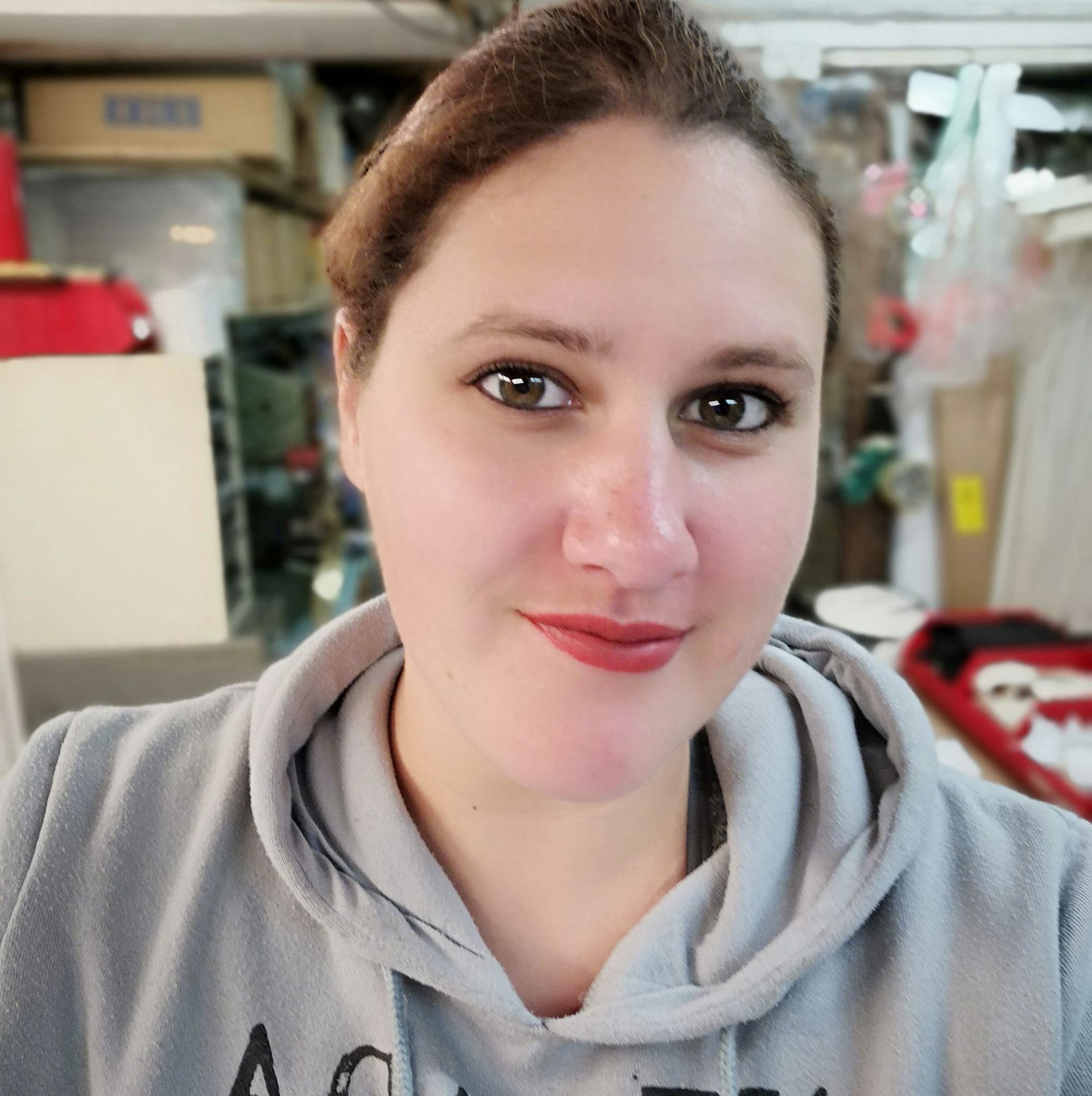 Vrouw in grijze hoodie lacht, bruin haar naar achteren gebonden, rode lippenstift, rommelige achtergrond.