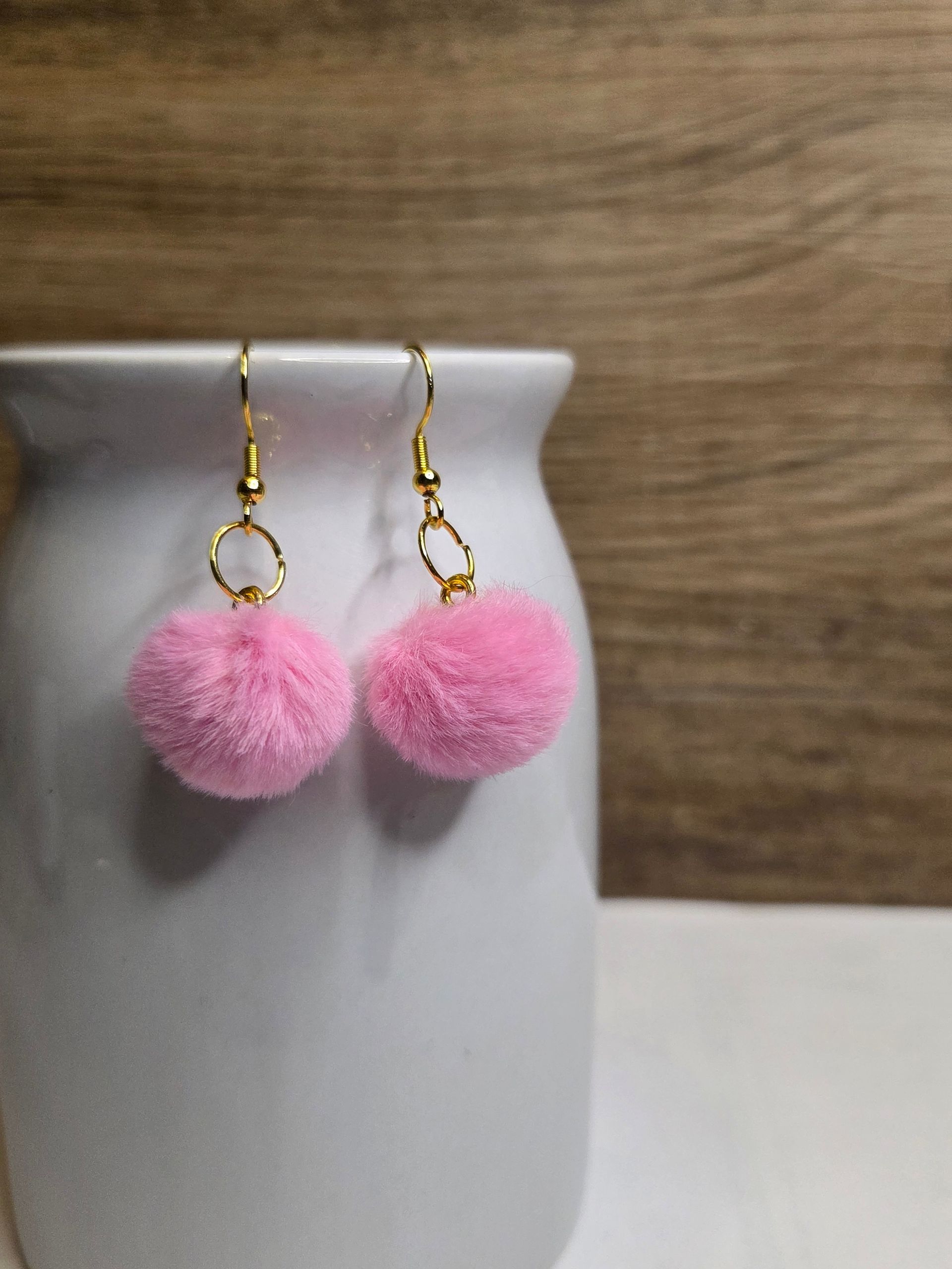 Roze pompon-oorbellen hangen aan gouden haakjes en staan ​​op een witte vaas tegen een houten achtergrond.